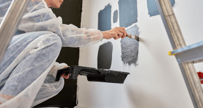 Persoon in beschermende overall verft een muur met grijstinten tijdens een doe-het-zelfproject thuis.